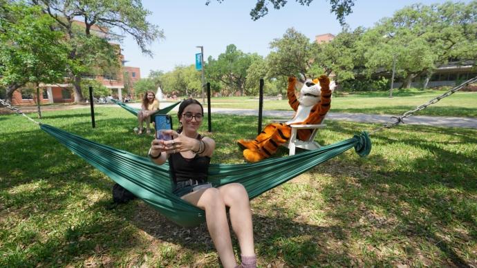 a girl sits in a green hammock taking a selfie with her phone. leeroy sits in adirondack chair behind her, and another girl is in a hammock behind her.