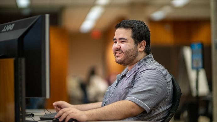 thomas pena sits at a desk in coates library, his hand on a computer mouse, while looking at a dell computer