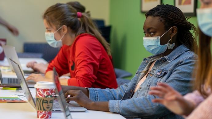 students wear masks and sit at desks while working on their laptops