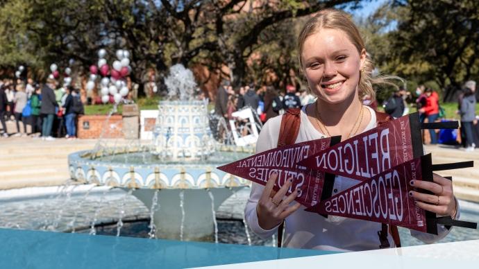 Student holding up degree pennants for Religion, 地球科学, and Environmental Studies in front of Miller fountain