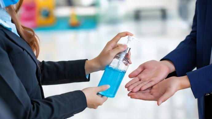 On right: pair of hands extended; on left: someone holds a bottle of hand sanitizer to put on the other person's hands