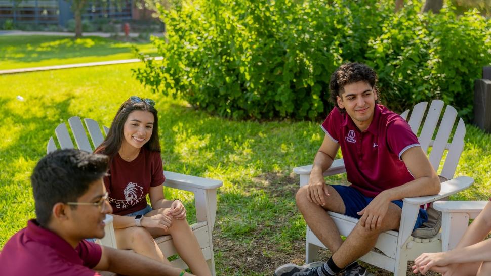 Students wearing Trinity shirts sit in Adirondack chair and chat outside