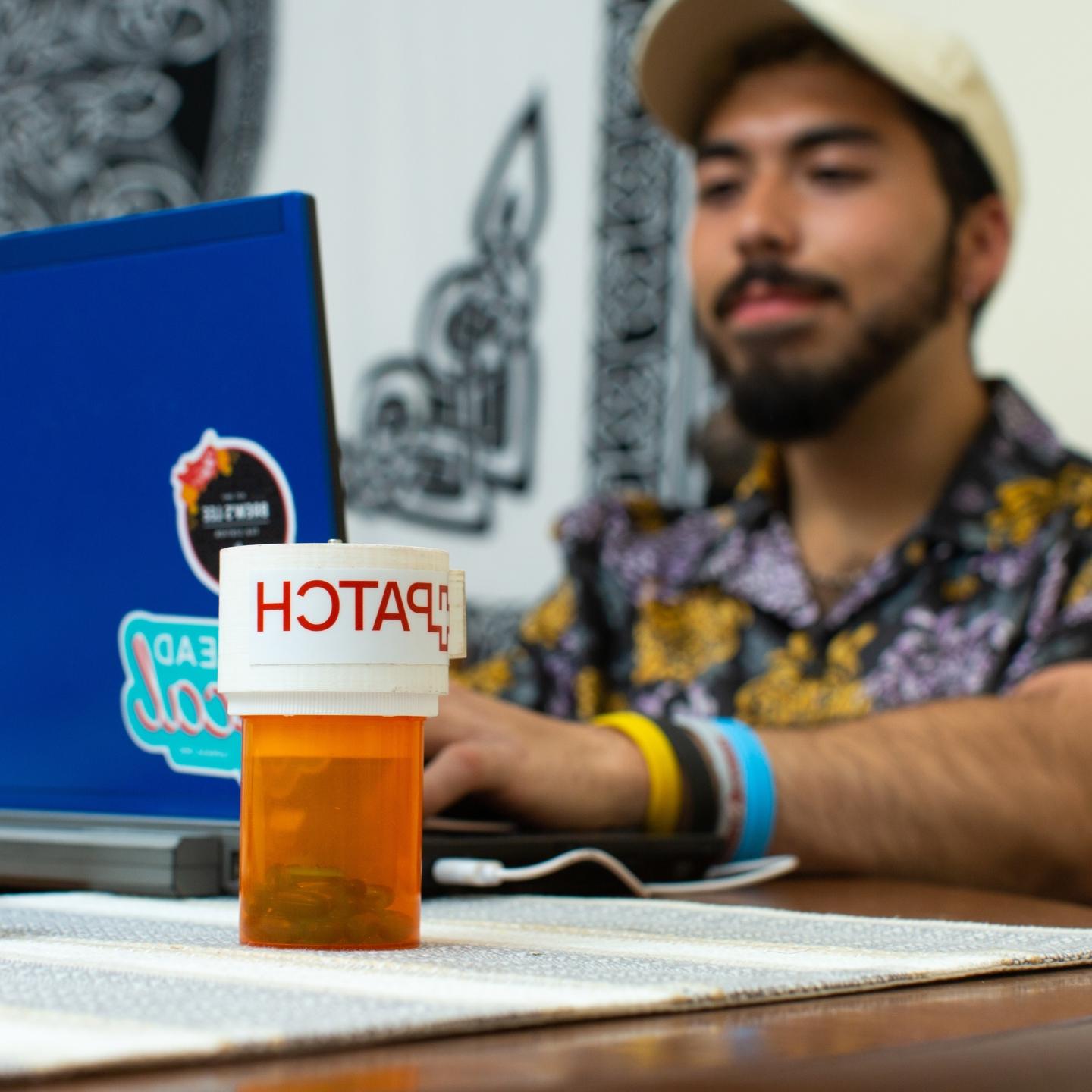 A PATCH pill bottle is in focus in the foreground, while Gavin Buchanan types on his laptop in the background.